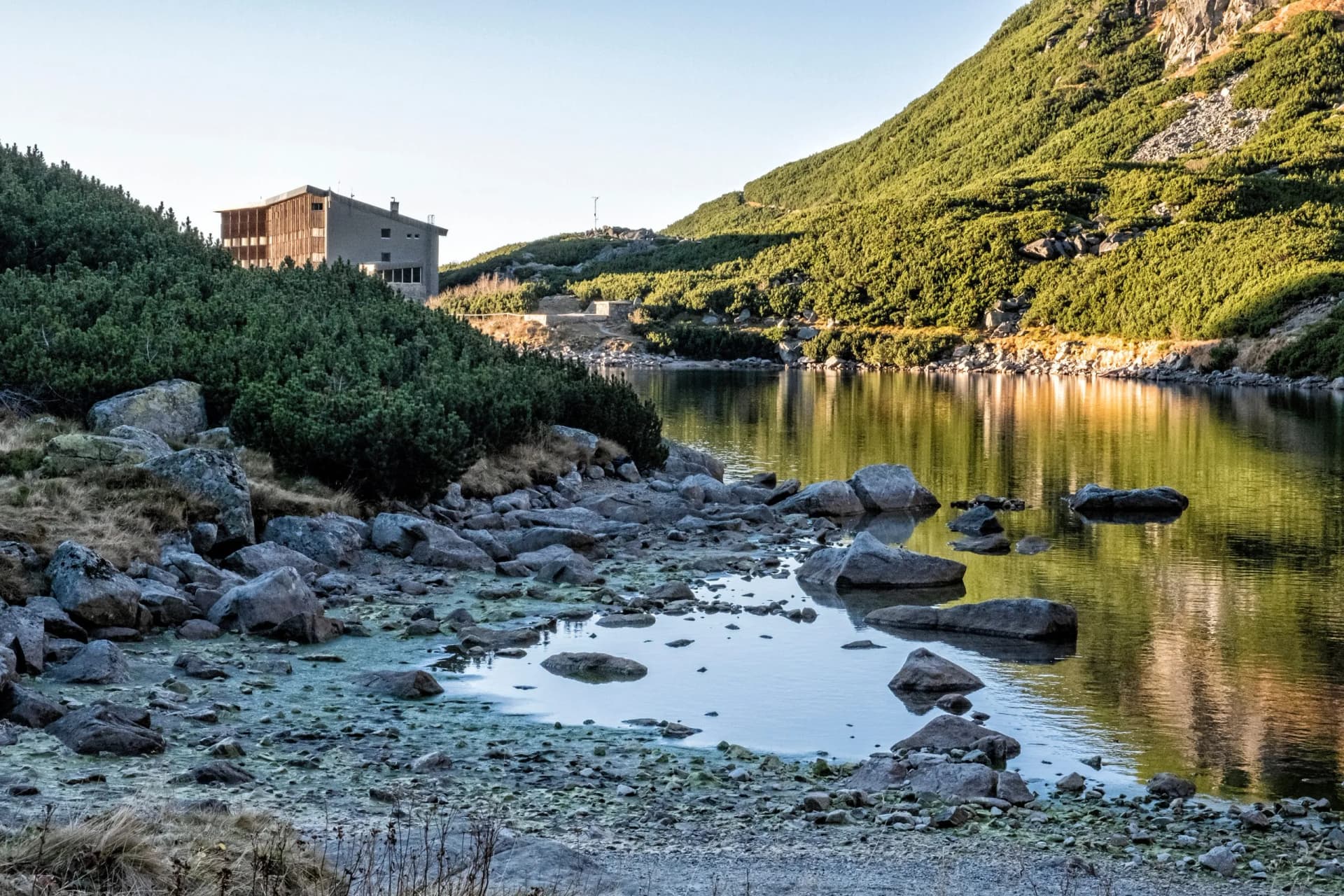 Mountain hut by rocky shore of alpine lake reflecting green hillside under clear sky, Sliezsky Dom.