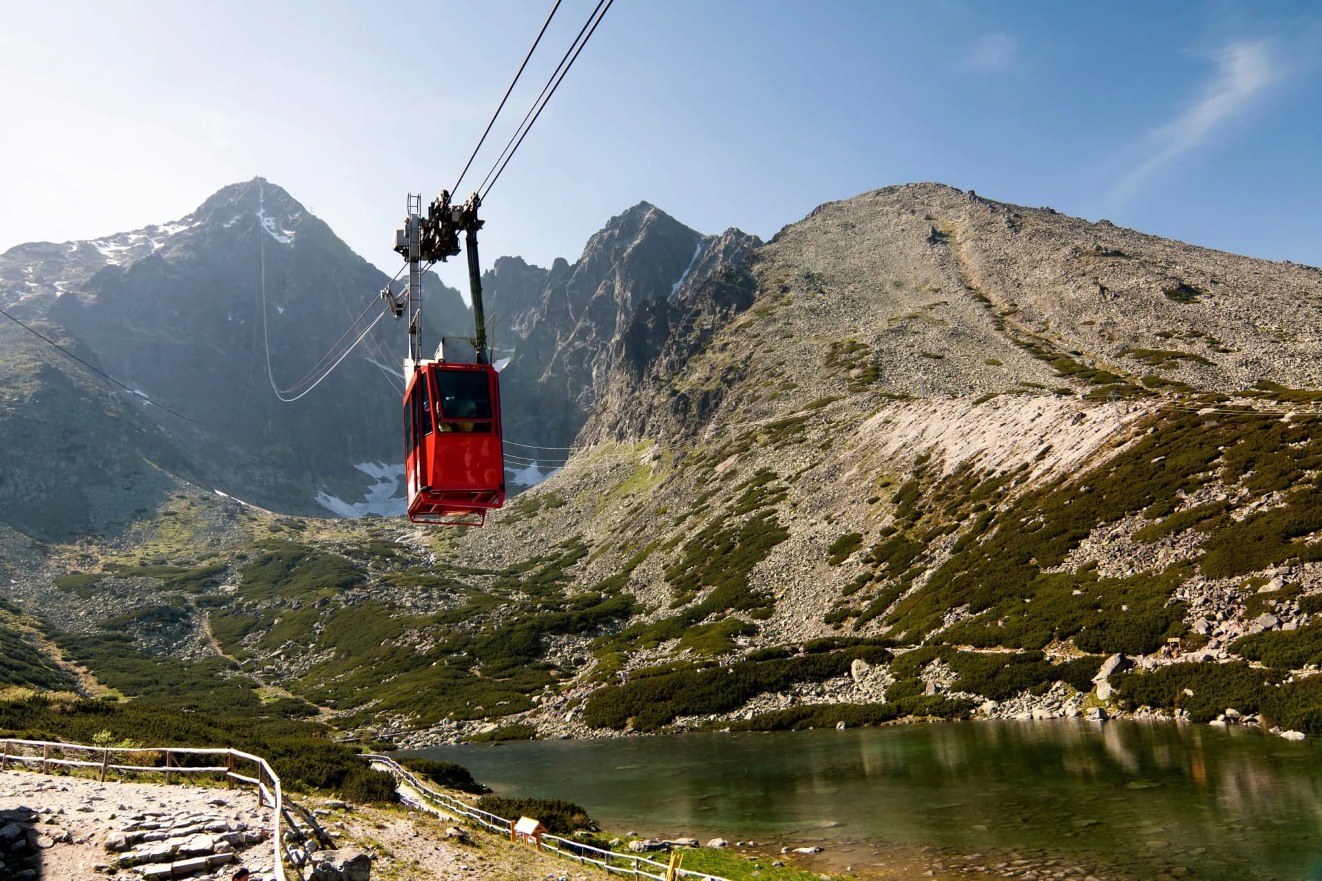 Red cable car ascending above Lomnicky Peak in the Tatras mountains near a clear lake.