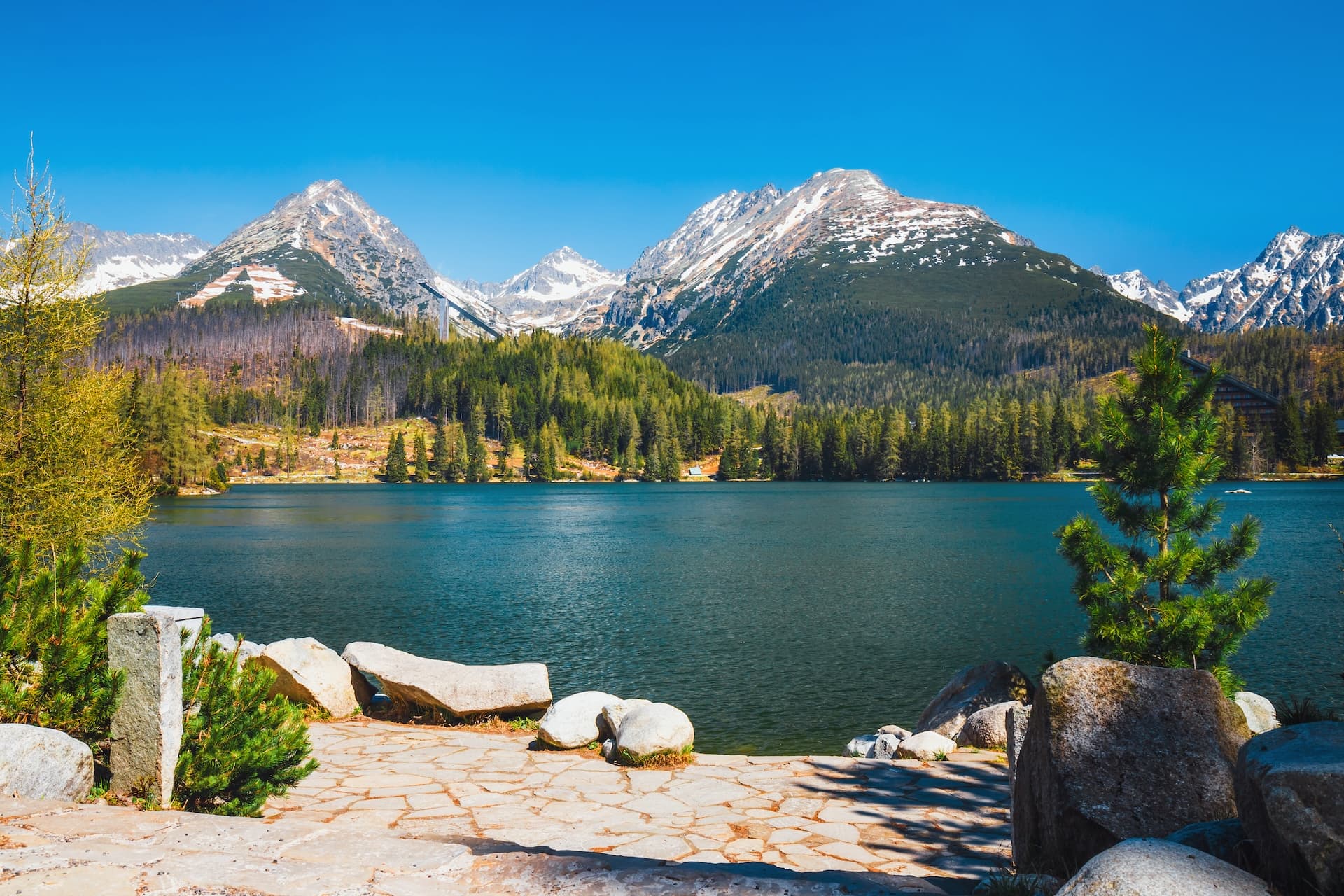 Mountain lake with snow-capped peaks, pine forest, and stone shoreline in the Tatra Mountains.