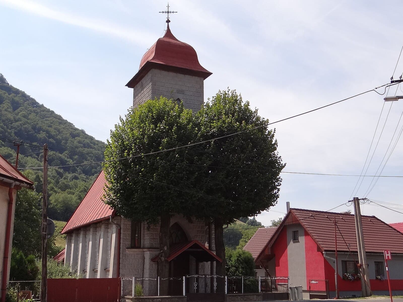 Church with red roof and stone tower in Kráľovany village against forested mountain backdrop.