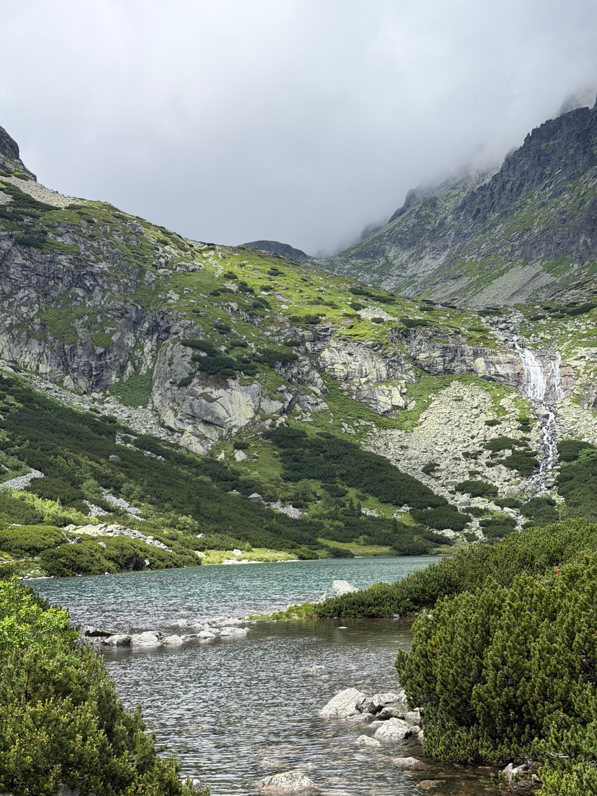 Alpine lake and waterfall in high mountain valley with green slopes and cloudy sky