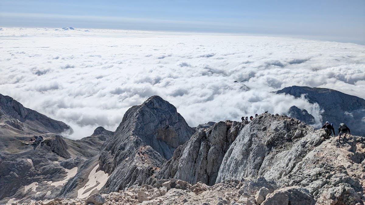 Hikers traversing rocky mountain ridge above sea of clouds under blue sky